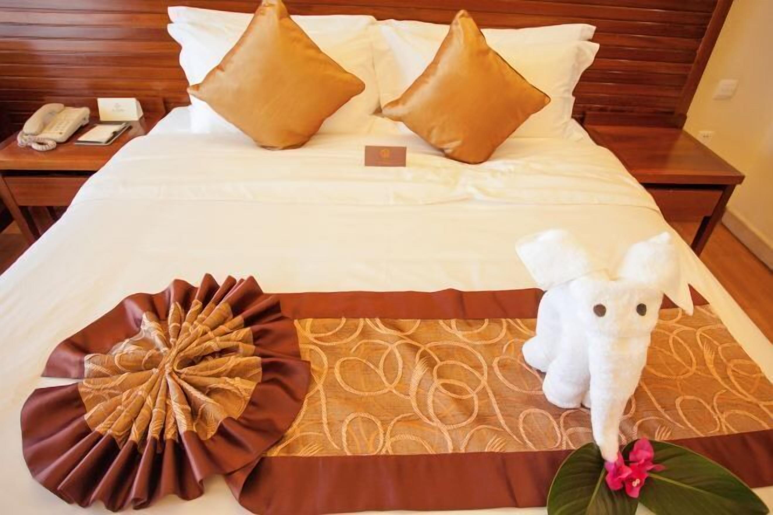 A neatly made bed with two pillows, a decorative brown and beige headboard, a white stuffed animal, and a small table with a telephone and a notepad.