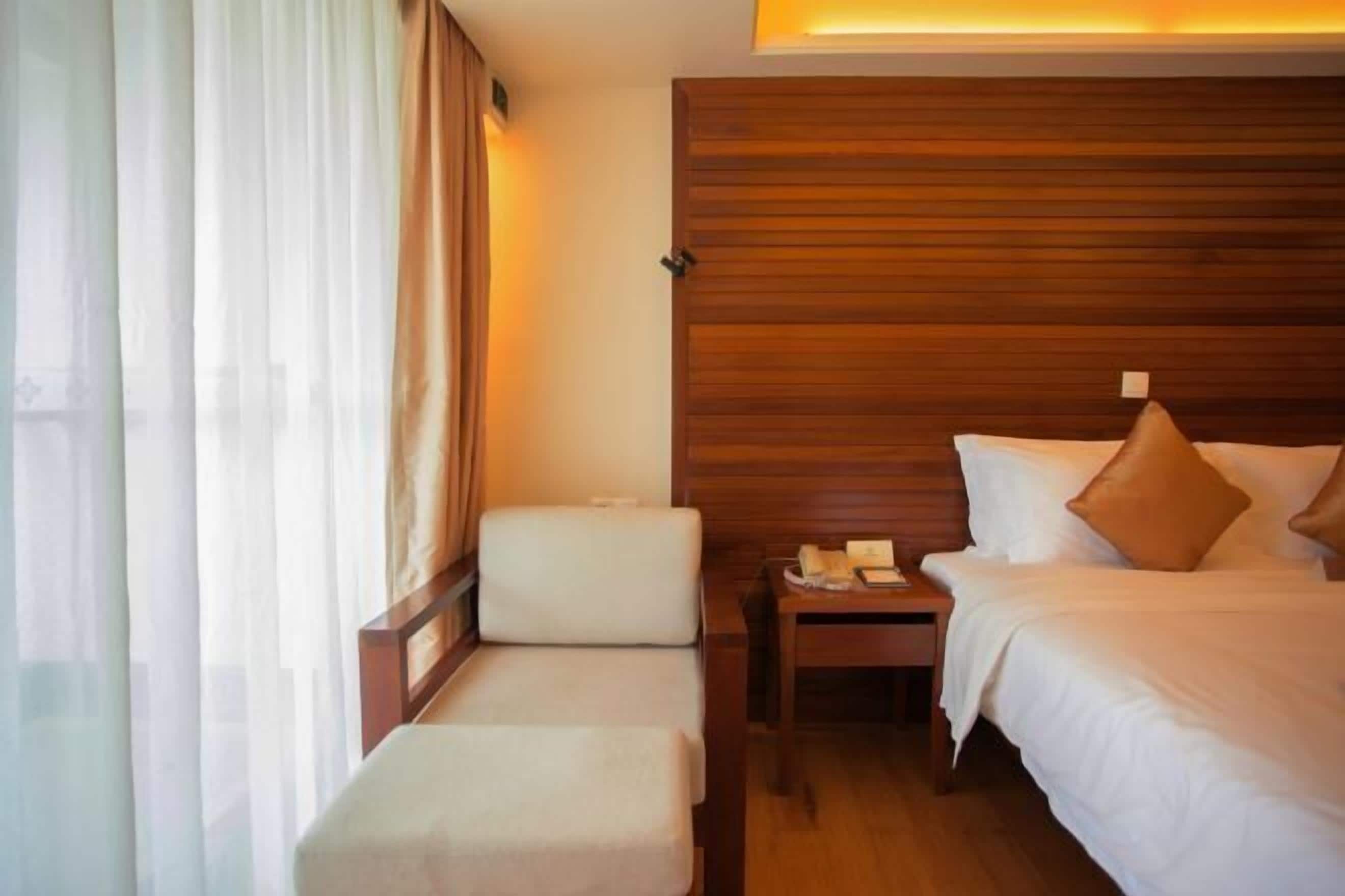 A hotel room with a wooden headboard, two beds, a bedside table with a lamp, a chair, and sheer curtains.
