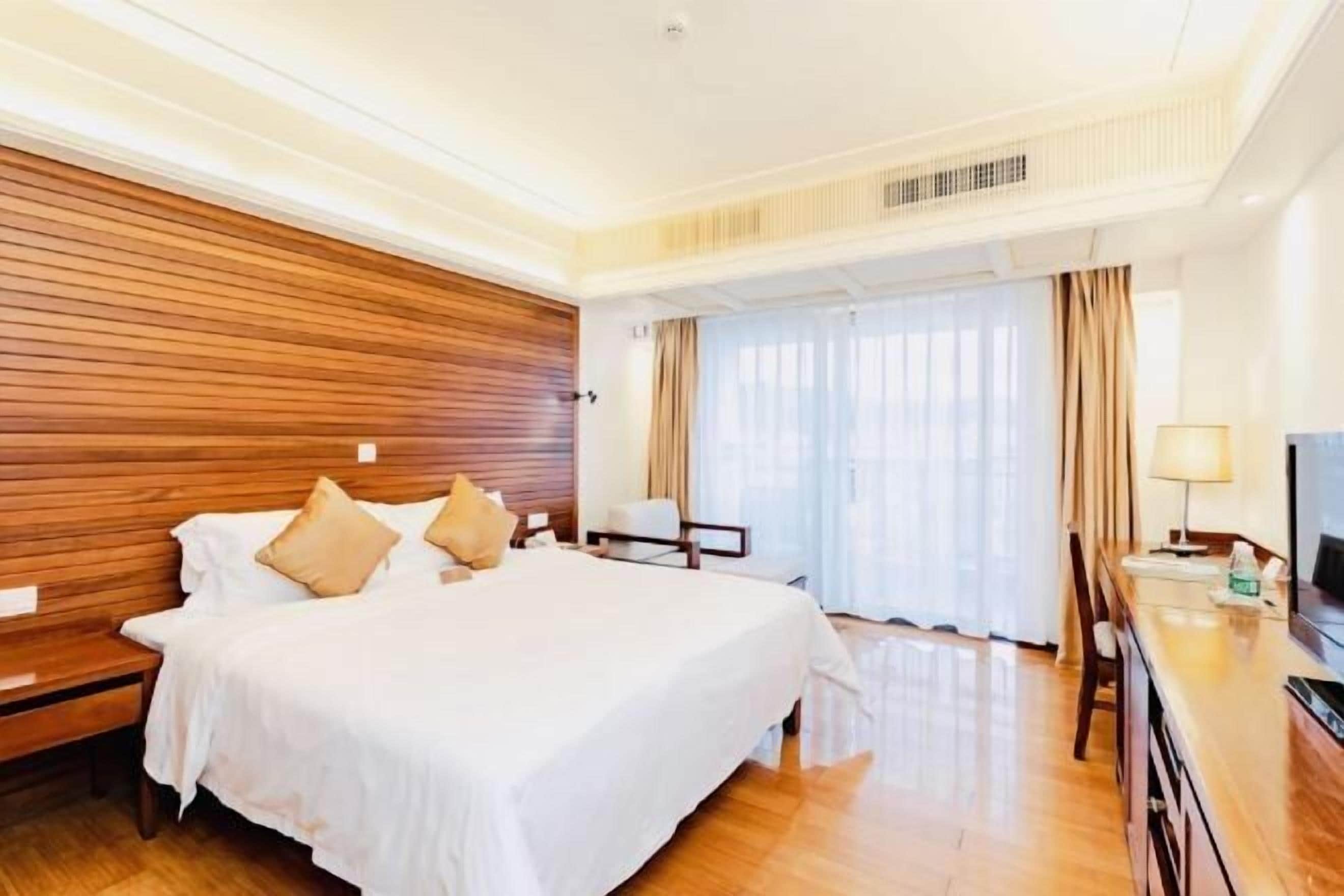 A hotel room with a wooden headboard, a large bed with white bedding, a desk with a lamp, and a television.