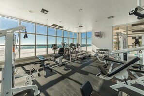 A gym with large windows overlooking a beach, various exercise equipment, and a TV mounted on the wall.