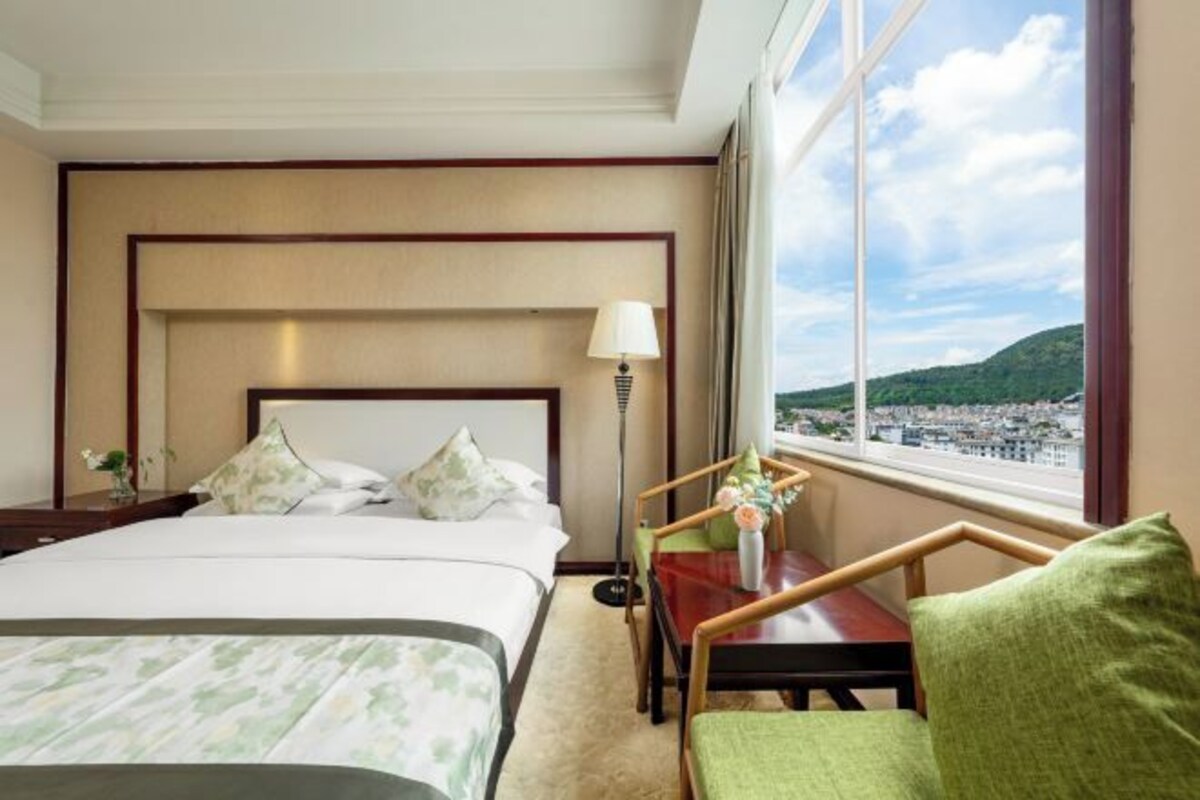 A hotel room with a bed, a desk, a chair, and a large window offering a city view.