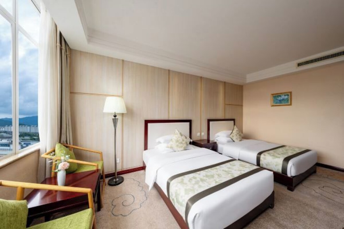 A hotel room with two beds, a desk, a chair, a lamp, and a view of the city.