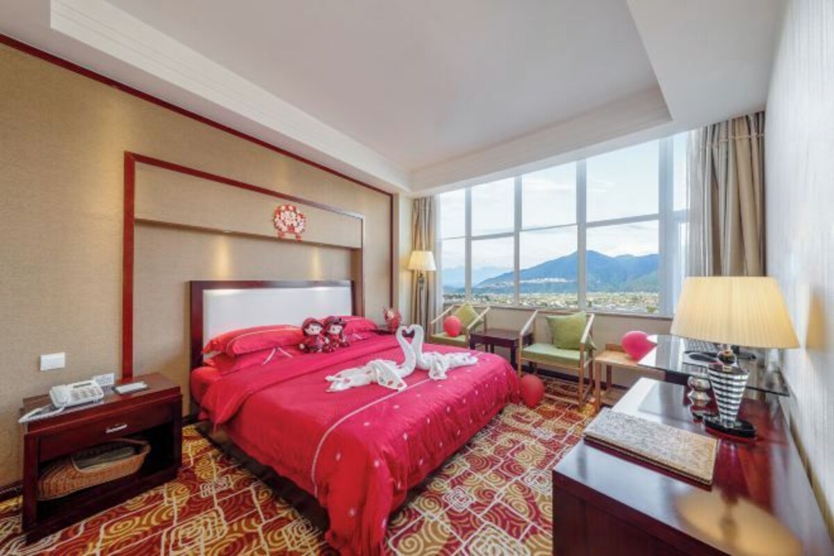 A hotel room with a large bed, a view of mountains, a desk with a lamp, and a chair.
