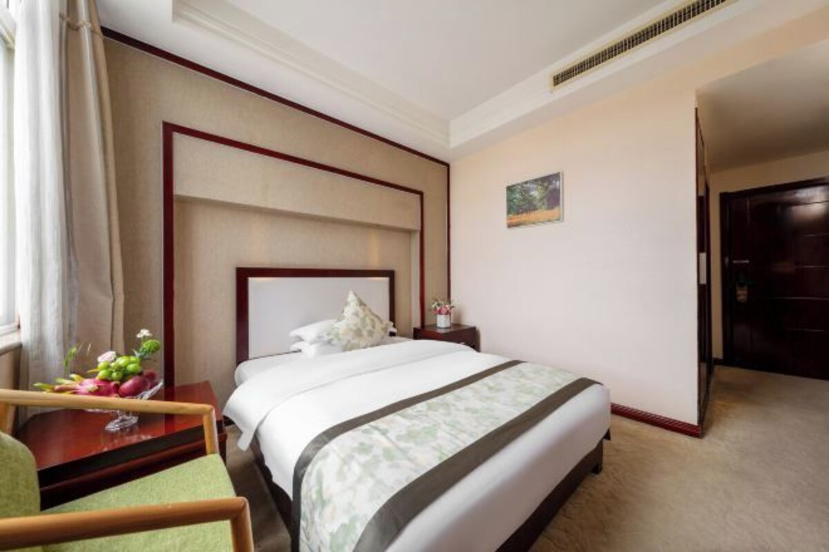A hotel room with a large bed, a bedside table with a vase of flowers, a chair, and a painting on the wall.