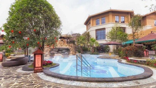 A hot spring area with a stone-tiled floor, a small tree with red flowers, and a building with large windows in the background. - Bailing Hot Spring Hotel (Anshun)