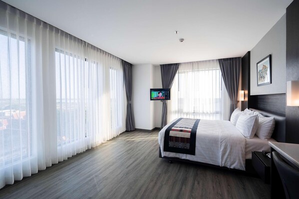 Ein Hotelzimmer mit einem großen Bett, einem Flachbildfernseher und Blick auf die Stadt durch durchsichtige Vorhänge.