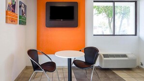 A room with an orange accent wall, a flat-screen TV mounted above a white table, two black chairs, and an air conditioning unit. - Magnuson Hotel Baton Rouge East (Baton Rouge)