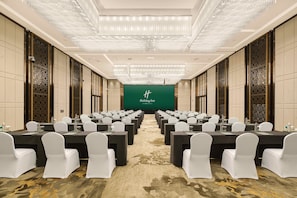 A conference room with rows of tables and chairs, a green screen with 'Holiday Inn' text, and a patterned carpet.