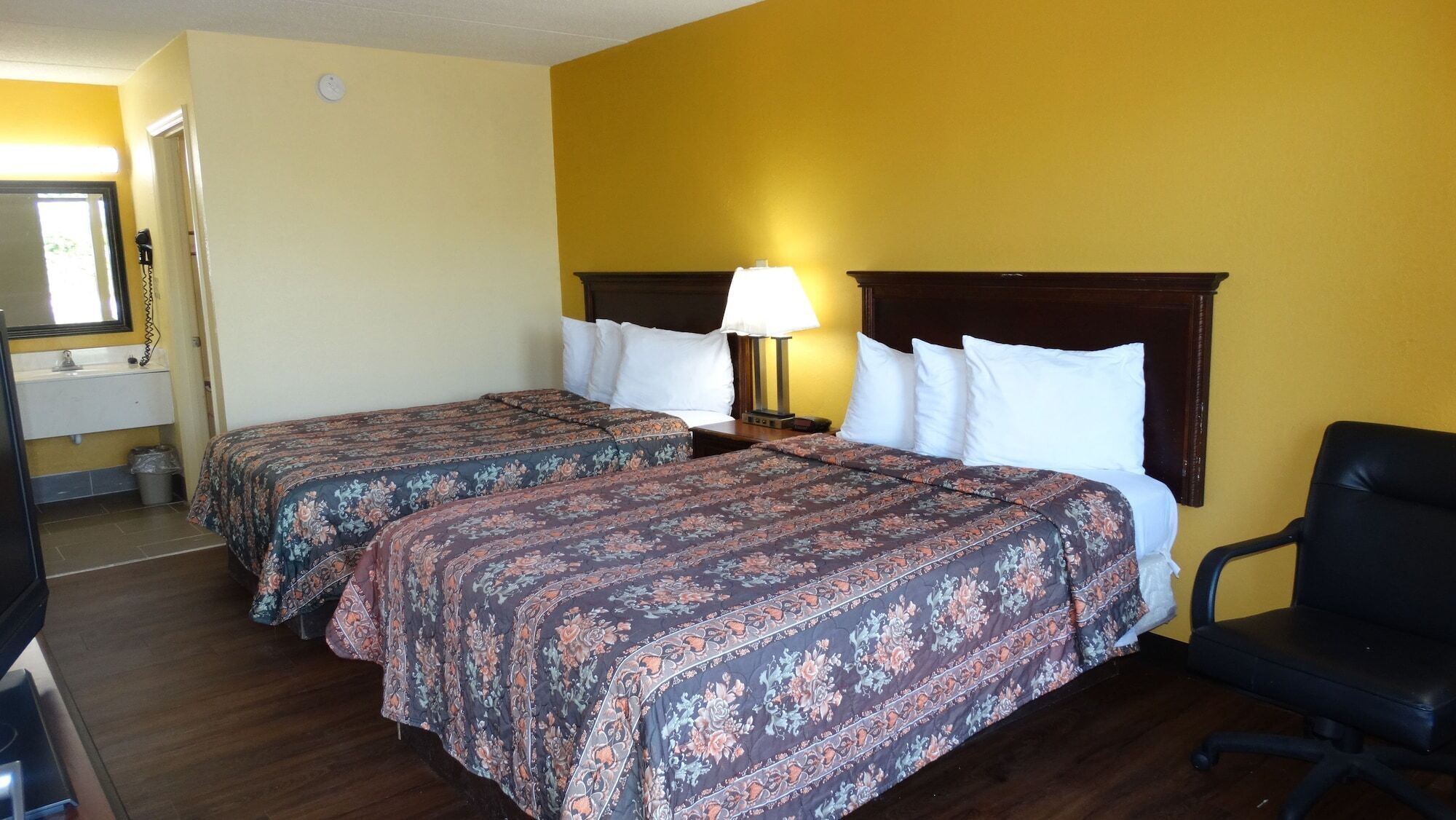 A hotel room with two beds, a nightstand with a lamp, a black office chair, and a bathroom with a sink and mirror.