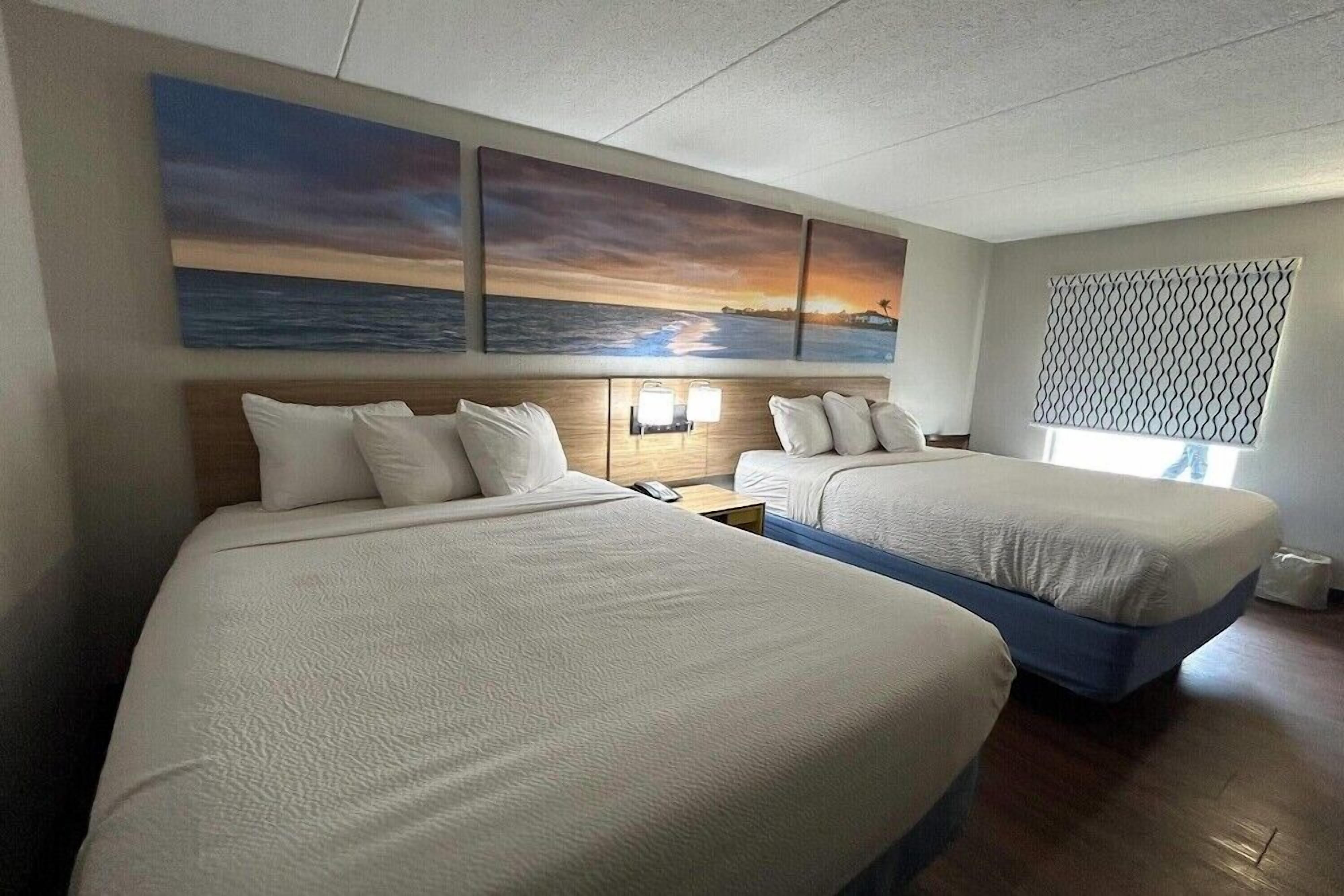Une chambre d’hôtel avec deux lits, une table de chevet avec une lampe, une fenêtre avec un store à motifs et un tableau représentant un coucher de soleil accroché au mur.