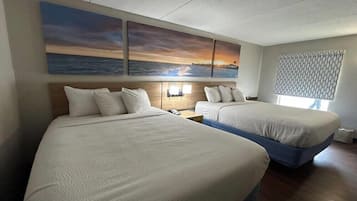 Une chambre d’hôtel avec deux lits, une table de chevet avec une lampe, une fenêtre avec un store à motifs et un tableau représentant un coucher de soleil accroché au mur.