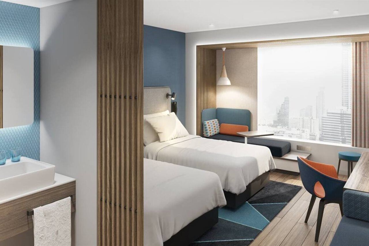 A modern hotel room with two beds, a large window with a city view, a desk with a chair, and a bathroom with a sink and mirror.