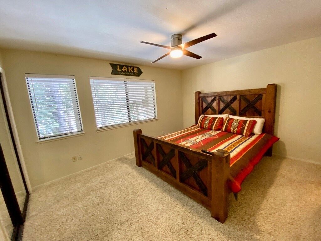 A bedroom with a wooden bed, two windows with blinds, a ceiling fan, and a sign on the wall.