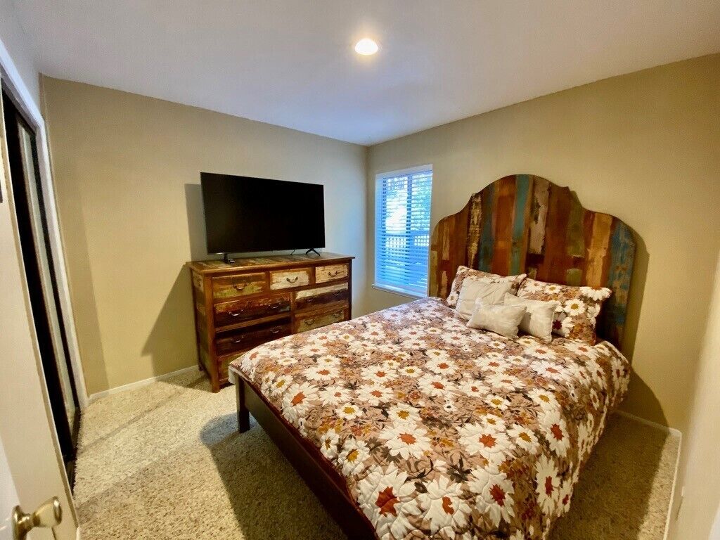 Een slaapkamer met een bed, een dressoir, een televisie en een raam met jaloezieën.