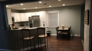 Cozinha moderna com ilha central, banquetas, mesa de jantar com cadeiras e janela com persianas.
