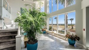 A spacious indoor area with a large potted plant, a staircase, and large windows offering a view of palm trees. - 2026 Updated, 5-Star, Modern Beachfront Condo at Silver Beach Towers (Destin)