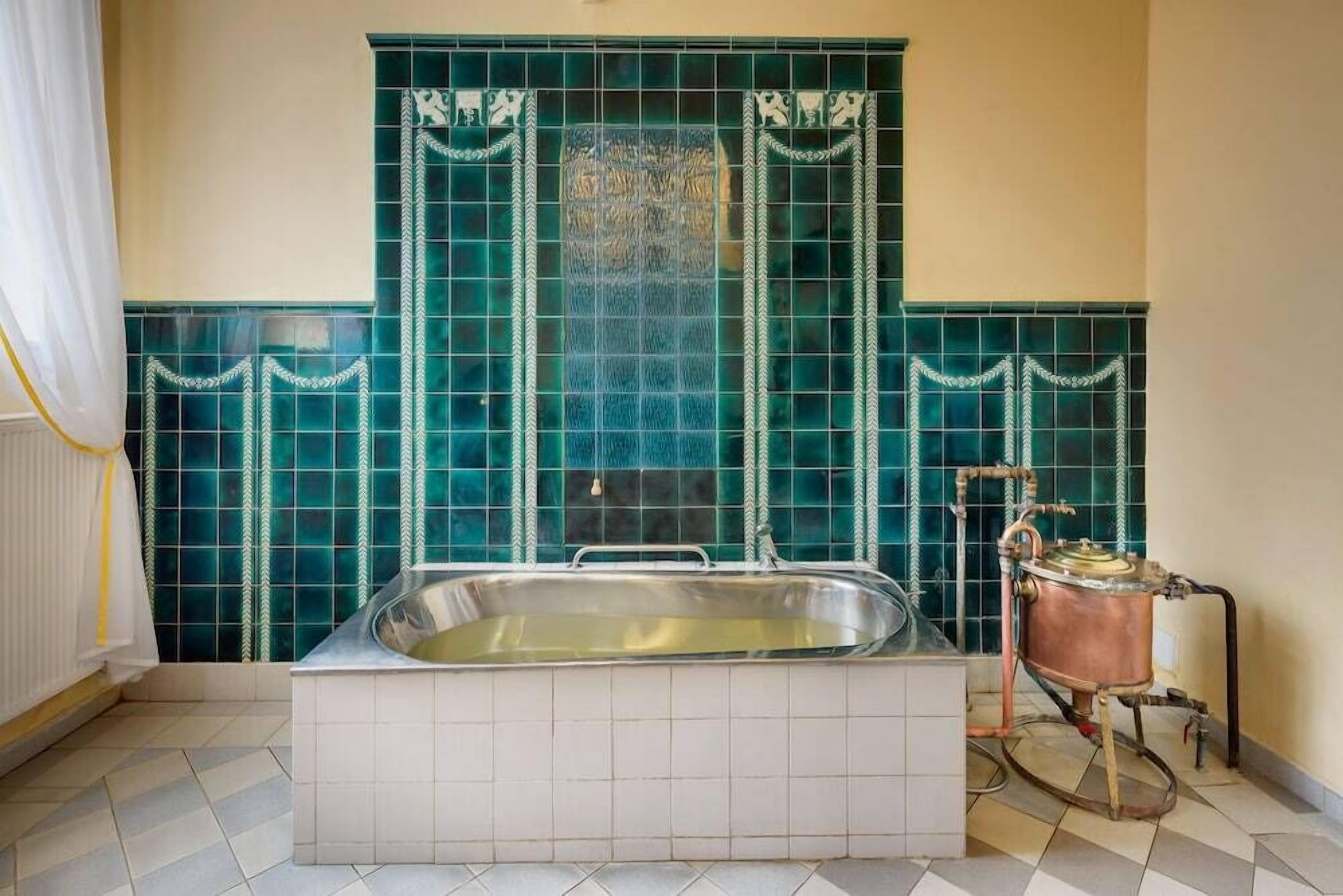 A bathroom with a bathtub, a copper water heater, and green-tiled walls.