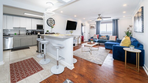 Une cuisine moderne avec des meubles blancs, un îlot central doté de tabourets, un téléviseur à écran plat et un ventilateur de plafond.