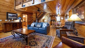 Un ampio interno di una baita in legno con soffitto alto, una scala, un soggiorno con divano blu, una zona pranzo e un'ampia finestra.
