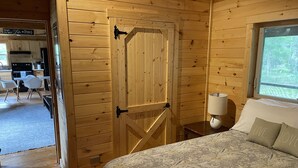 Puinen mökki sisältä, jossa on sänky, ikkunasta avautuva näkymä, keittiö sekä ruokapöytä ja tuolit.