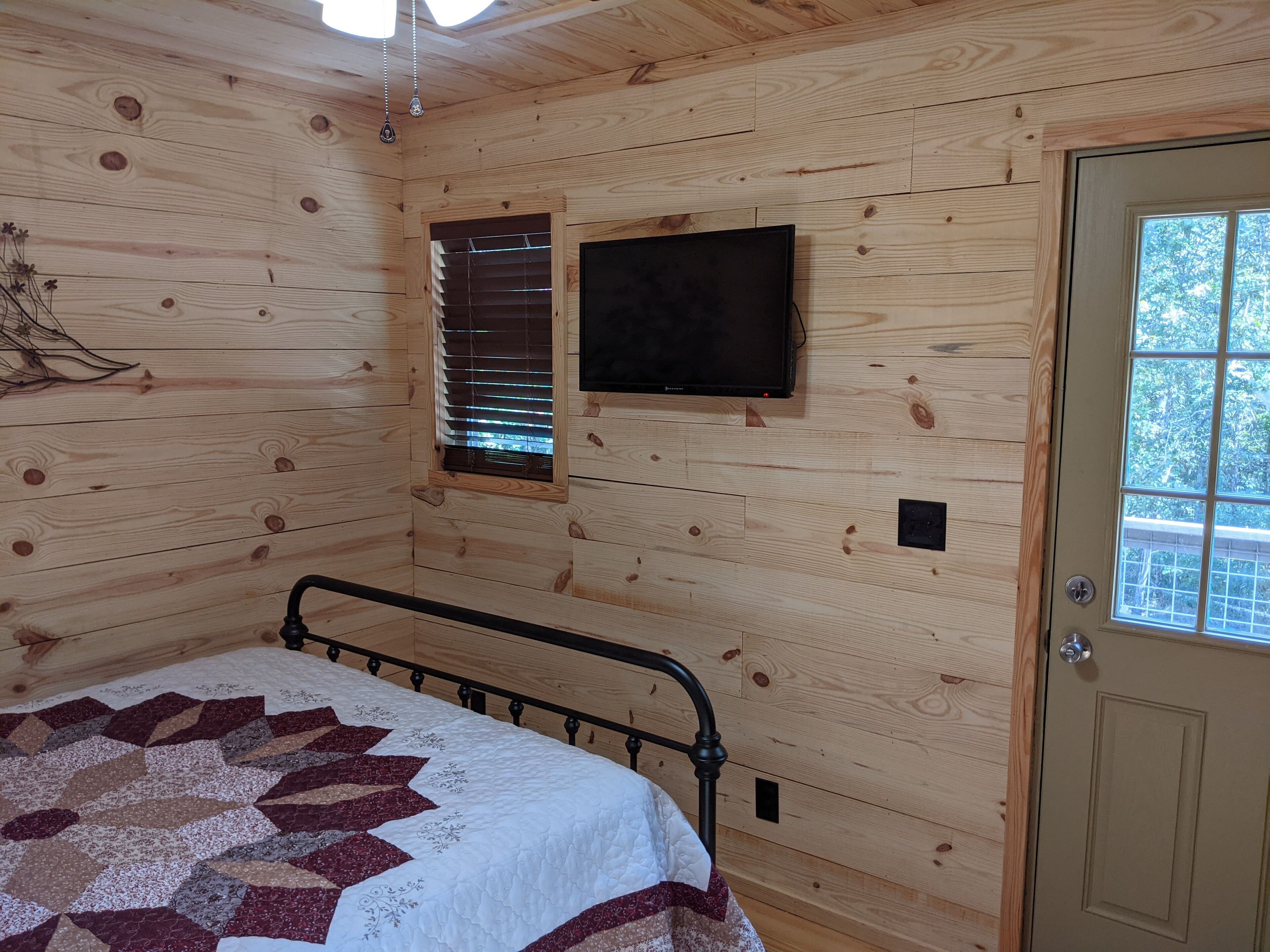A wooden cabin room with a bed, a window with blinds, a door, and a mounted television.