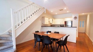 A dining area with a wooden table and black chairs, a kitchen with white cabinets, and a staircase with white railings. - Adelaide | 3BR Norwood Townhouse | Fringe Stay + Wi-Fi + Parking + Smart TV | Walk Everywhere (Norwood)