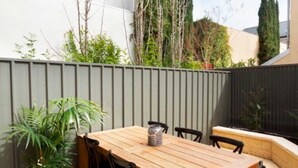 A patio with a wooden table and chairs, surrounded by a low fence and greenery. - Adelaide | 3BR Norwood Townhouse | Fringe Stay + Wi-Fi + Parking + Smart TV | Walk Everywhere (Norwood)