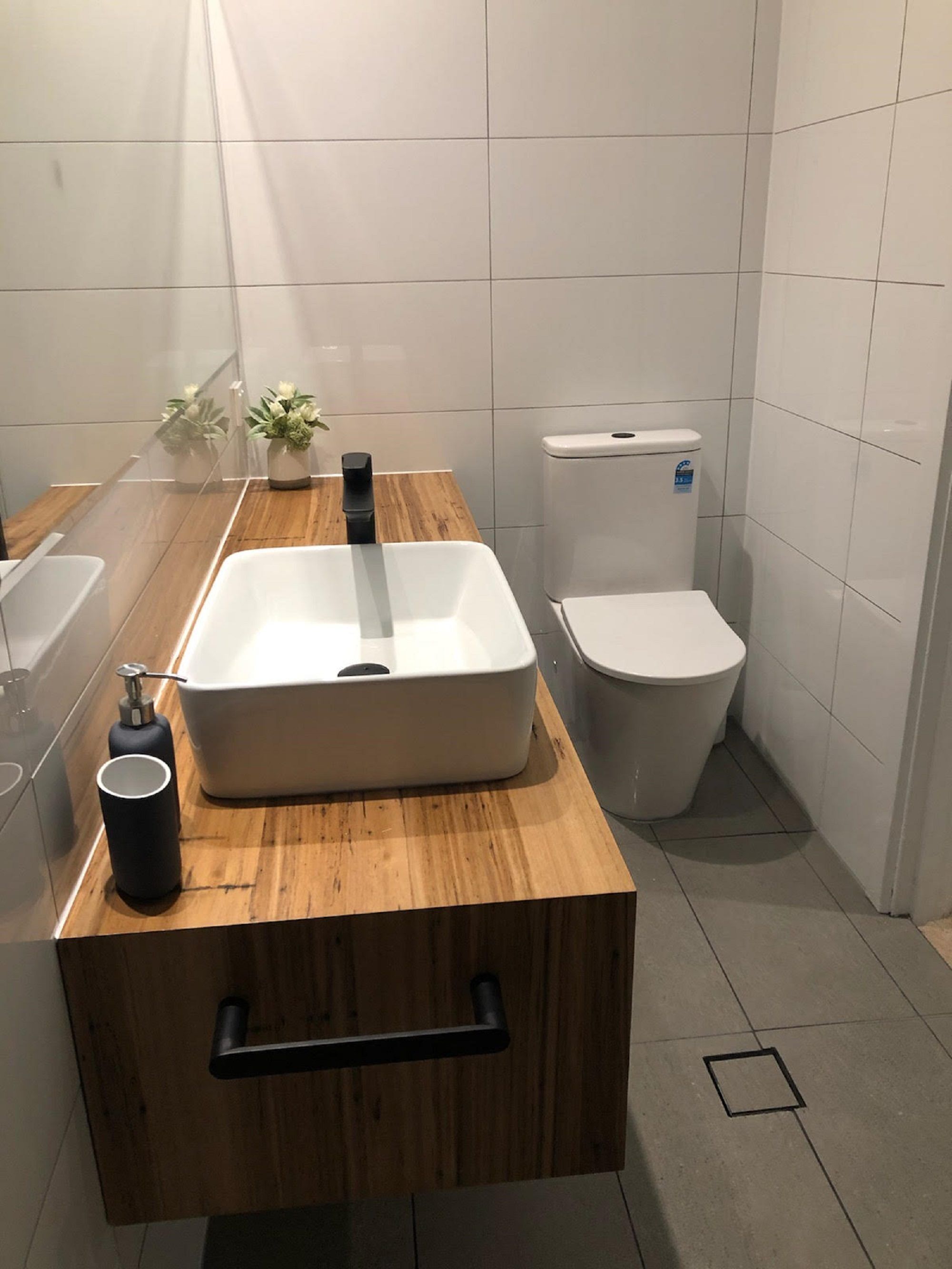 Um banheiro moderno com um espelho sobre bancada de madeira, pia branca, vaso sanitário e uma pequena planta em vaso.