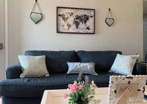 A living room with a dark blue sofa, two heart-shaped wall decorations, a world map, and a 'Home' pillow. - Convenient to Wind Farms, Wedding Venue, Hunting, Large Yard-Pet Friendly! (Lahoma)