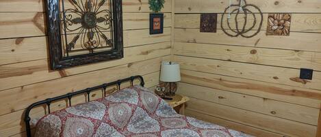A room with a bed, a ceiling fan, a wall-mounted picture frame, a small table with a lamp, and a wall decoration.