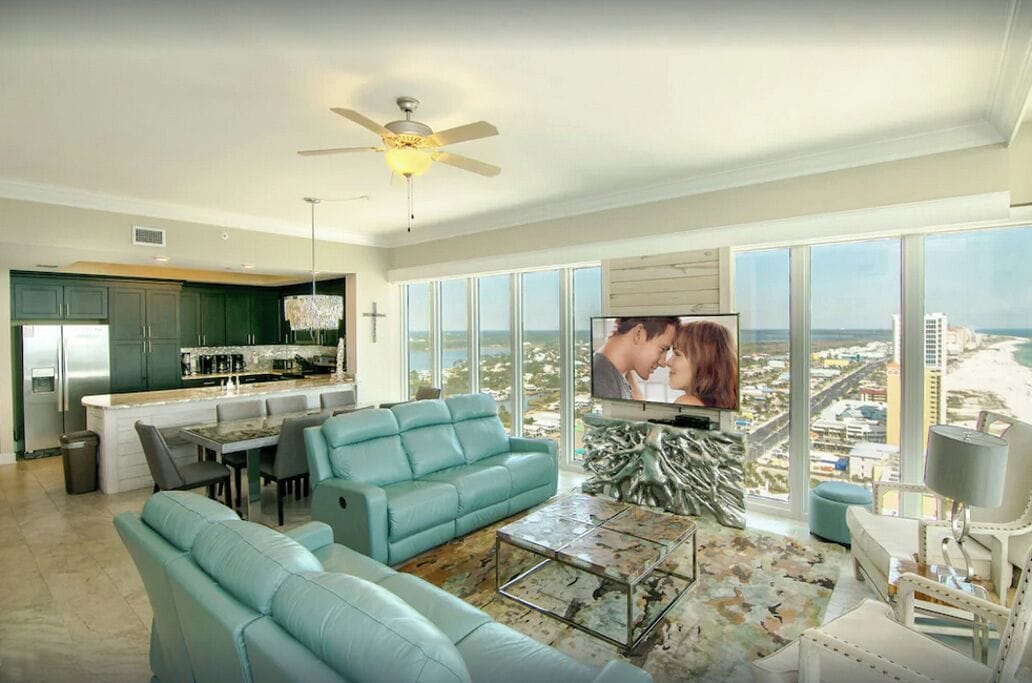 Una sala de estar moderna con sofá, mesa de centro y televisor, una cocina con barra, y ventanas grandes con vista a la ciudad.