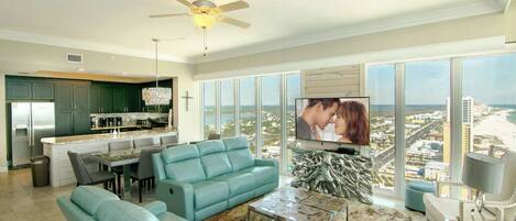 Una sala de estar moderna con sofá, mesa de centro y televisor, una cocina con barra, y ventanas grandes con vista a la ciudad.