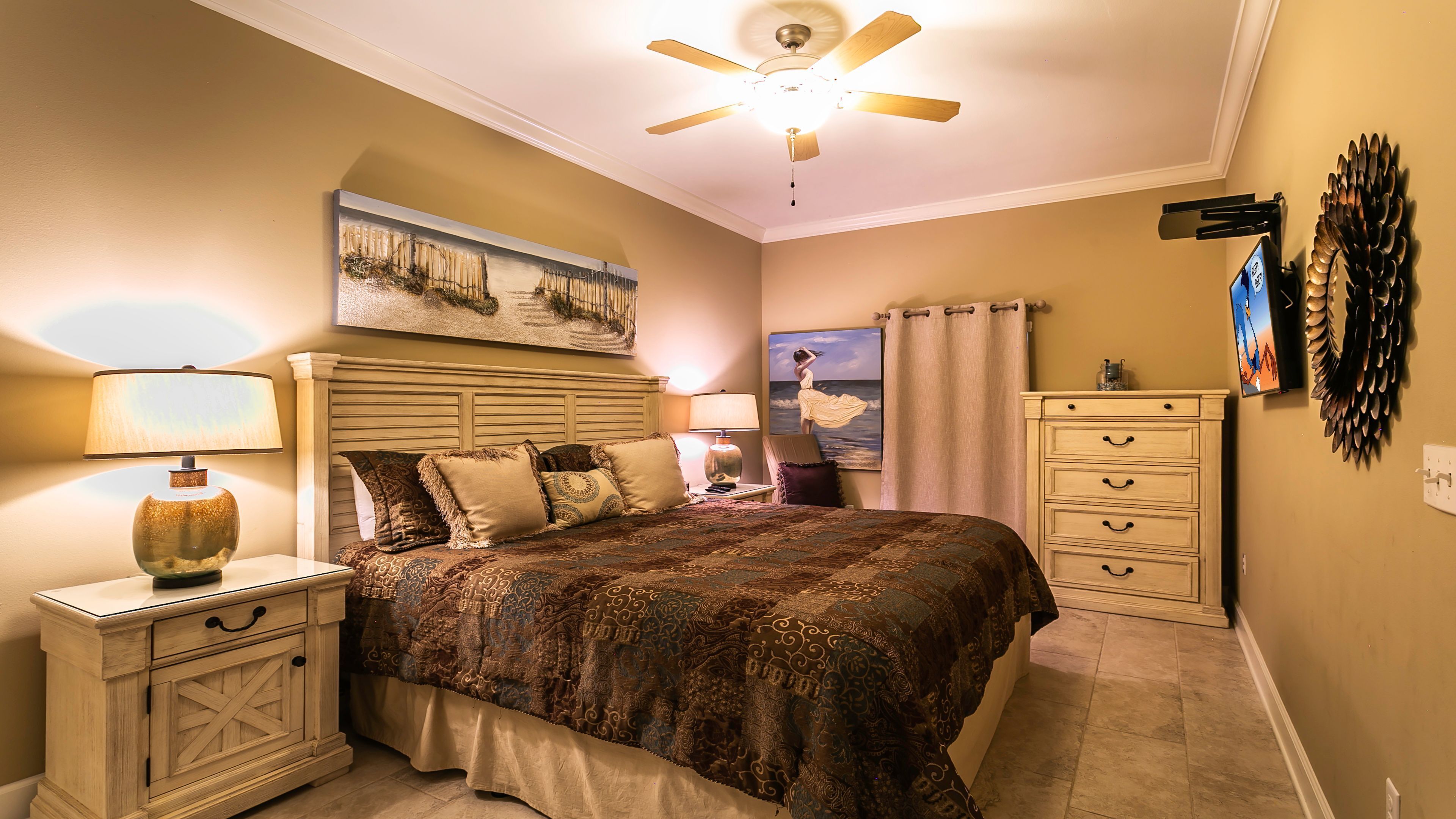 Une chambre à coucher comprenant un lit, deux tables de chevet, un ventilateur de plafond, un tableau, un téléviseur et un élément décoratif mural.