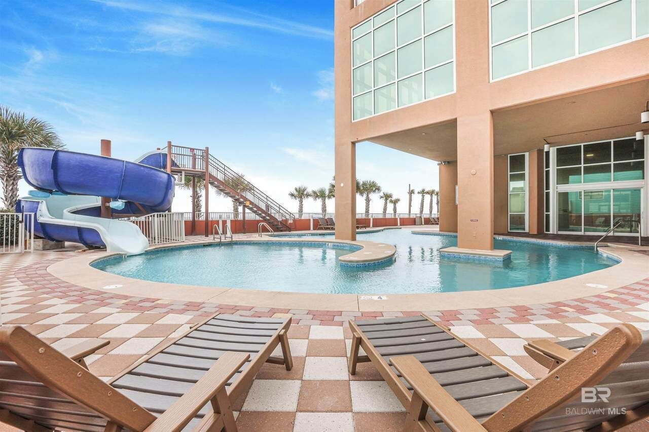A pool area with lounge chairs, a water slide, and a building with large windows.