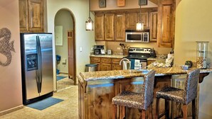 Una cocina con gabinetes de madera, electrodomésticos de acero inoxidable, encimera de granito y taburetes de mimbre.