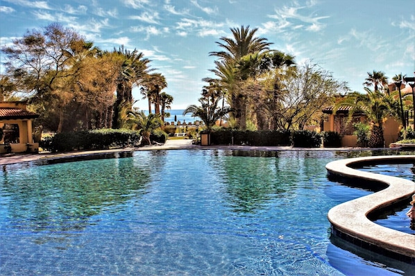 A clear pool with a curved edge, surrounded by palm trees and a building with a tiled roof.