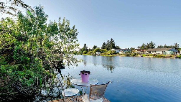 Een houten terras met metalen stoelen en een kleine tafel, uitkijkend op een rustig meer met huizen op de achtergrond.