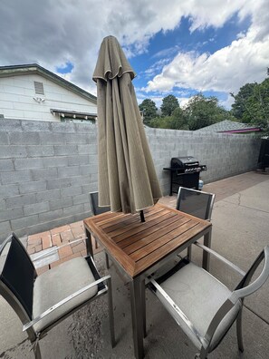 Een terras buiten met een houten tafel, vier stoelen, een parasol en een barbecue.