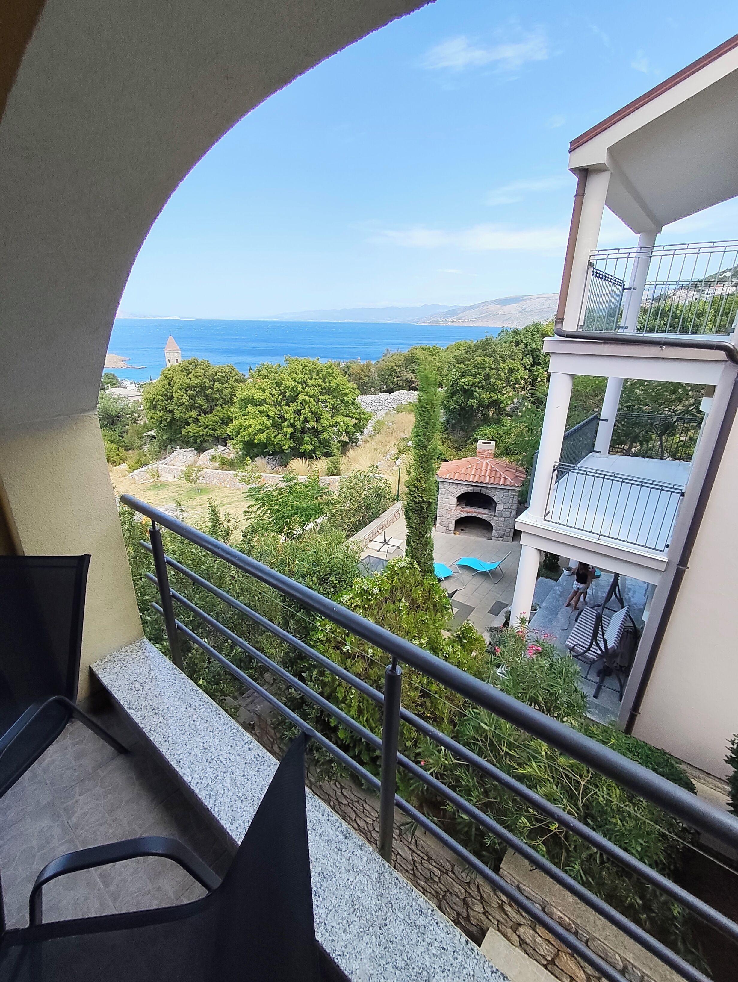 A balcony with a view of the sea, a stone structure, and a garden area.