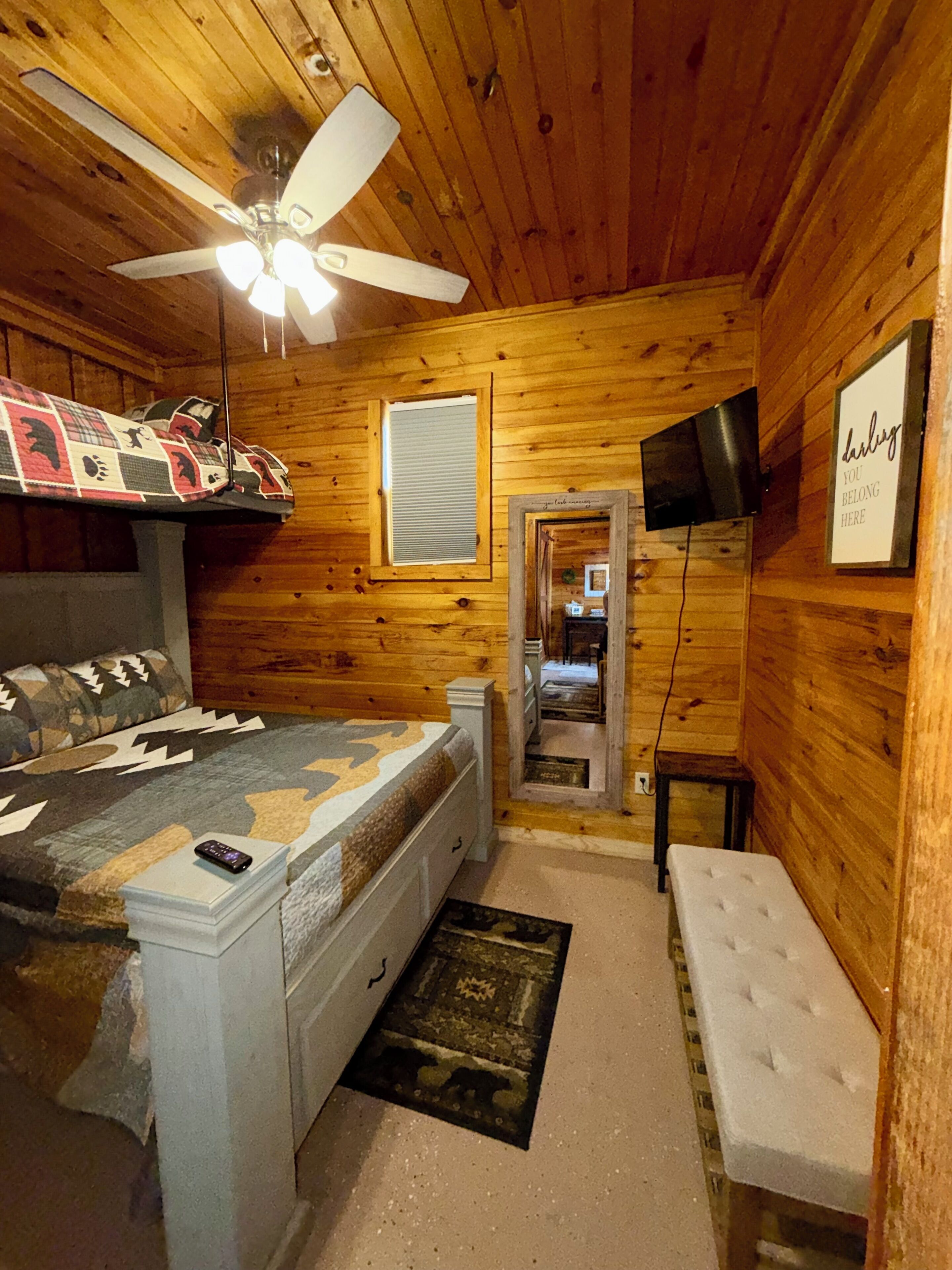 Une pièce de style cabane en bois, comprenant un lit, un banc, un ventilateur de plafond, une télévision et un miroir.