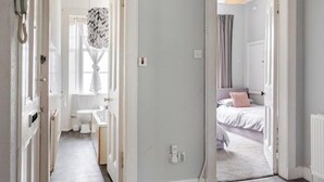 A narrow hallway leading to a bathroom with a toilet and sink, and a bedroom with a bed and curtains. - Our Wee Getaway (Rothesay)