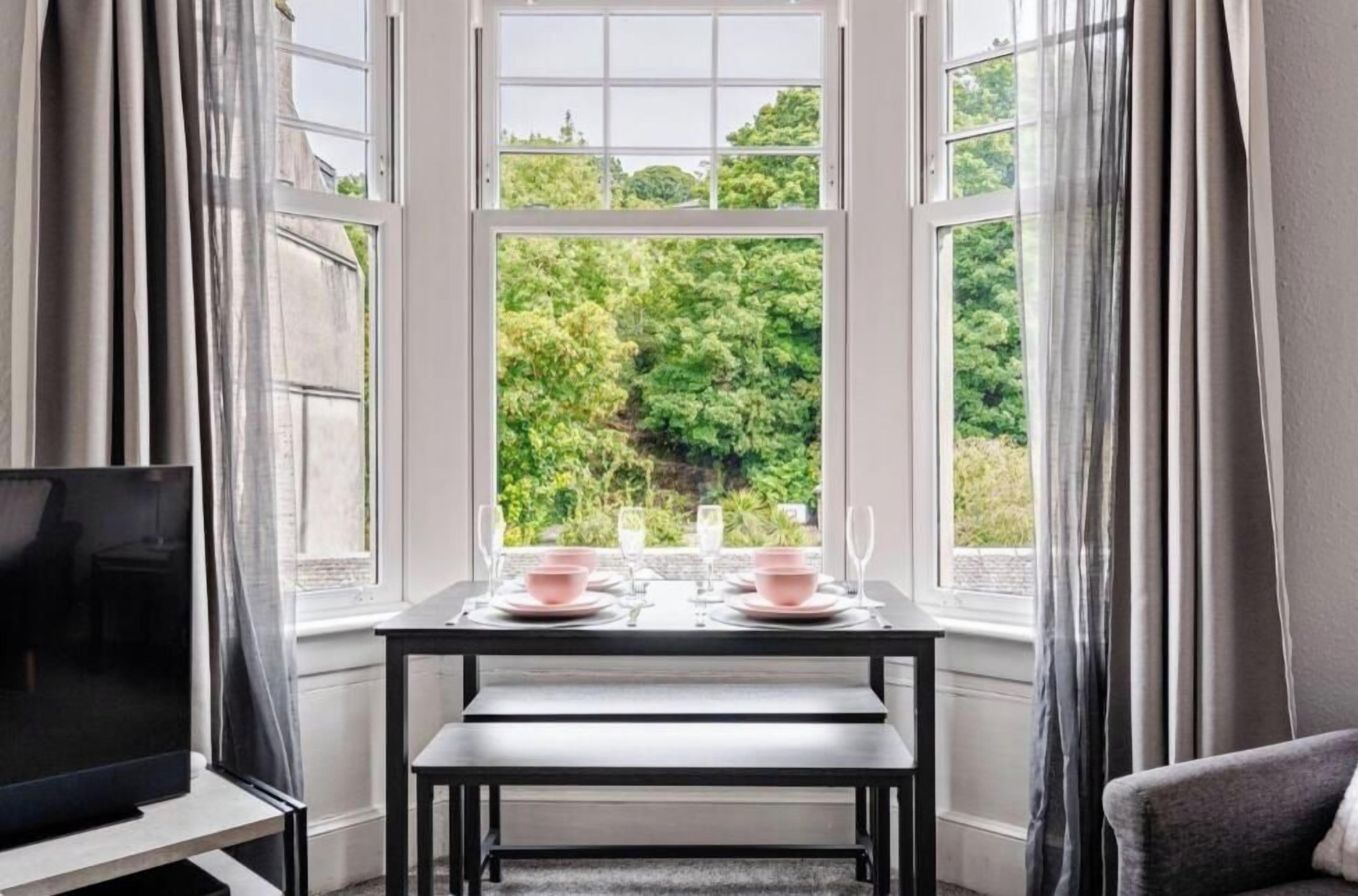 Un comedor con una mesa dispuesta para dos, un televisor sobre un mueble y vistas a la vegetación a través de amplios ventanales.
