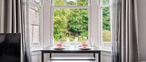 Un comedor con una mesa dispuesta para dos, un televisor sobre un mueble y vistas a la vegetación a través de amplios ventanales.
