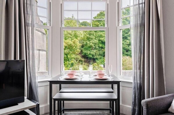 A dining area with a table set for two, a television on a stand, and a view of greenery through large windows.