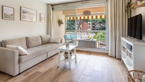 En moderne stue med en beige sofa, et lille hvidt bord og et fjernsyn på et hvidt stativ. En stor skydedør fører ud til en balkon med en pool og træer.