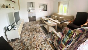 Un salon avec un canapé, un fauteuil, une table basse et un téléviseur. On aperçoit une cuisine au fond.