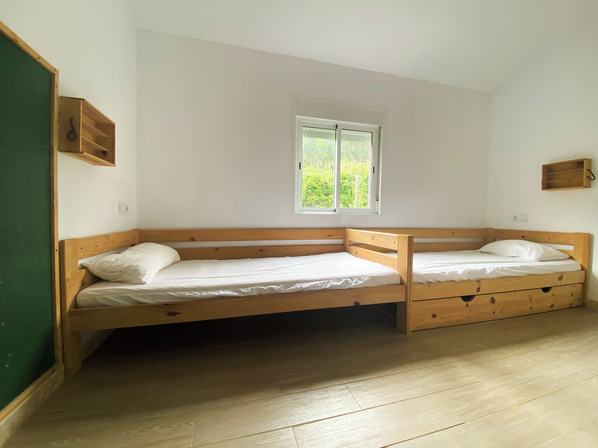 A room with two wooden bunk beds, a window with a view of greenery, and a small wooden shelf on the wall.