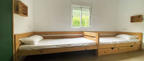 A room with two wooden bunk beds, a window with a view of greenery, and a small wooden shelf on the wall.
