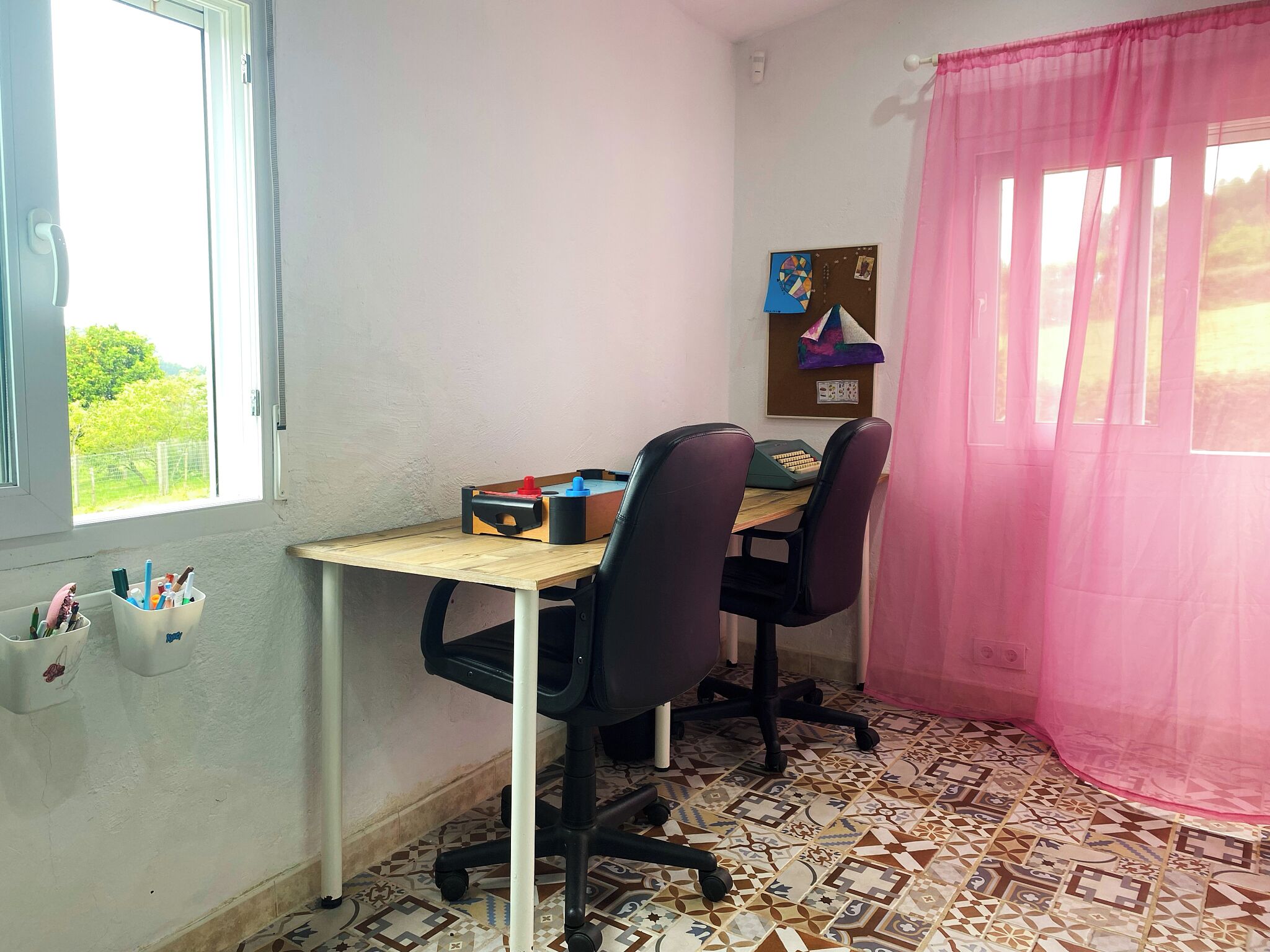 Una habitación con escritorio, dos sillas, ventanal con vistas, tablón de anuncios y suelo estampado.