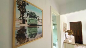 Un couloir avec un tableau encadré représentant une voiture ancienne, dont le reflet se mire dans une flaque d’eau, une porte en bois et une petite table sur laquelle est posé un vase.